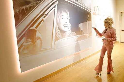 Dolly Parton in a pink suit pointing at a large black-and-white photograph of herself