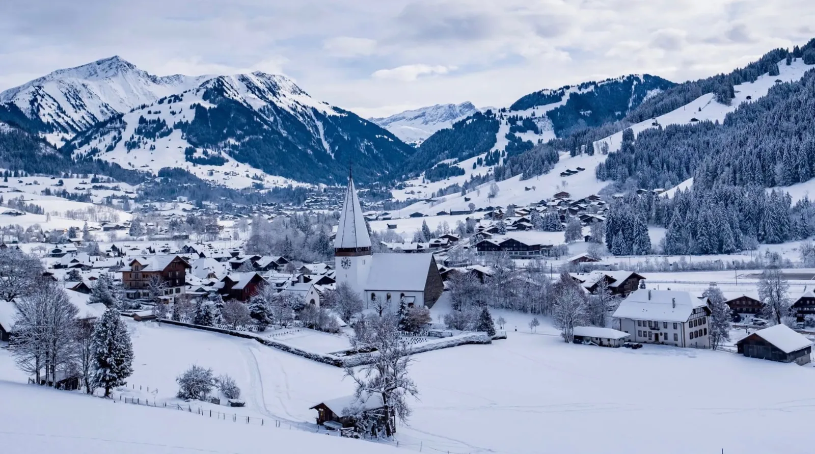 A Saanenland village under snow, its church spire rising among chalet roofs with mountains beyond
